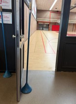 Foto van klapdeuren die leiden naar de sportzaal. De rechterklapdeur is dicht. De linkerklapdeur is opengezet met een zware paal. De doorgang is smal.